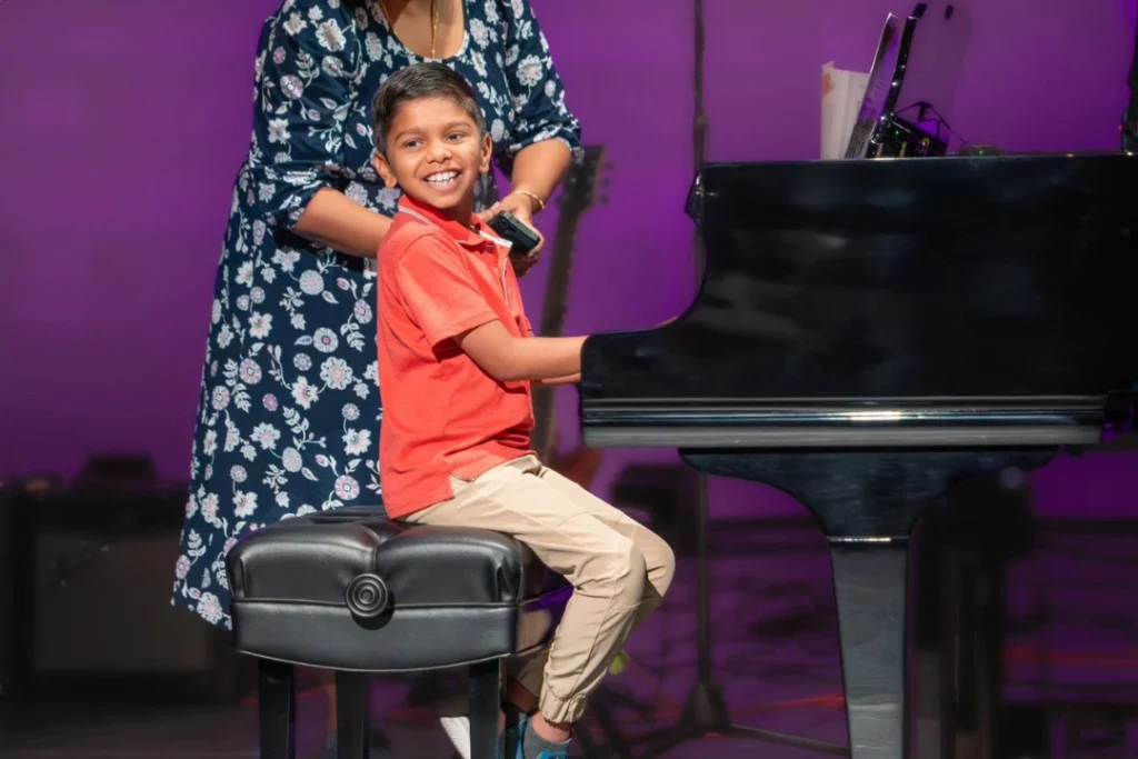 Smiling child playing piano confidently during a music recital at Gilbert School of Music, with an audience watching in the background